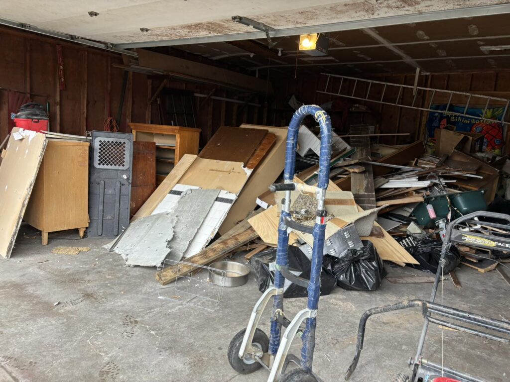 A cluttered garage filled with discarded furniture, wood scraps, and trash bags, highlighting a cleanout project.